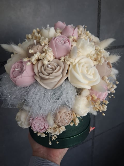 Hand holding a decorative flower arrangement with pink and cream roses, babys breath, and dried grasses in a green container