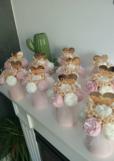 Collection of decorative floral arrangements with gold heart toppers, pink and white flowers, and beige textured bases on a white shelf with cactus in background