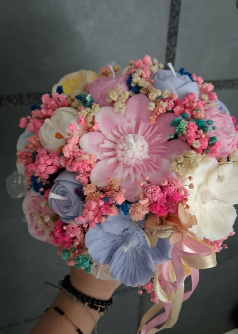 Hand holding a colorful bouquet of pink carnations, white and blue flowers with pink ribbon against a gray background