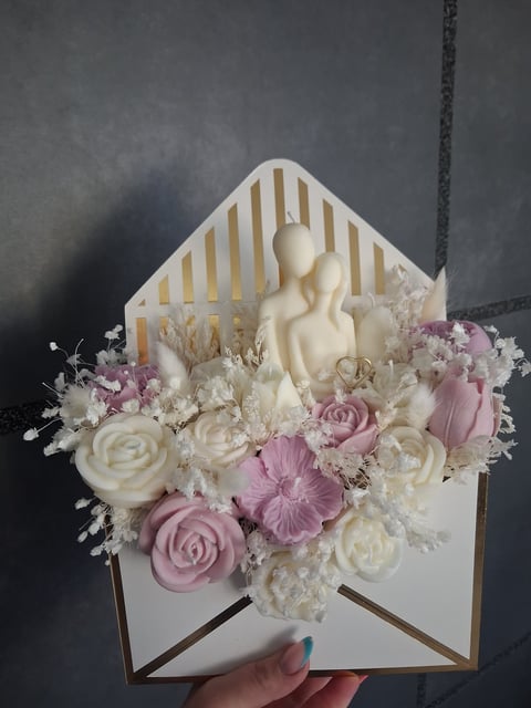 Decorative open envelope with cream and pink flowers, white ceramic figure, and dried botanicals arranged inside against a striped backdrop